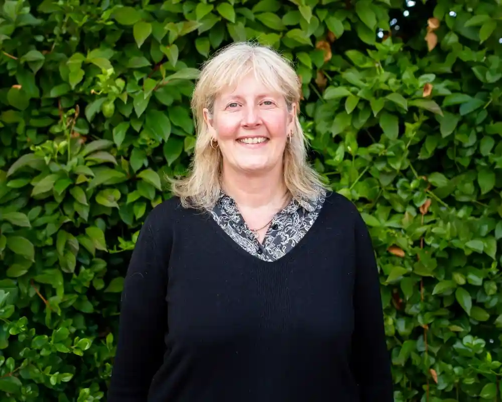 A smiling woman in a black sweater stands in front of a lush green hedge, with her hair partly wavy and light.