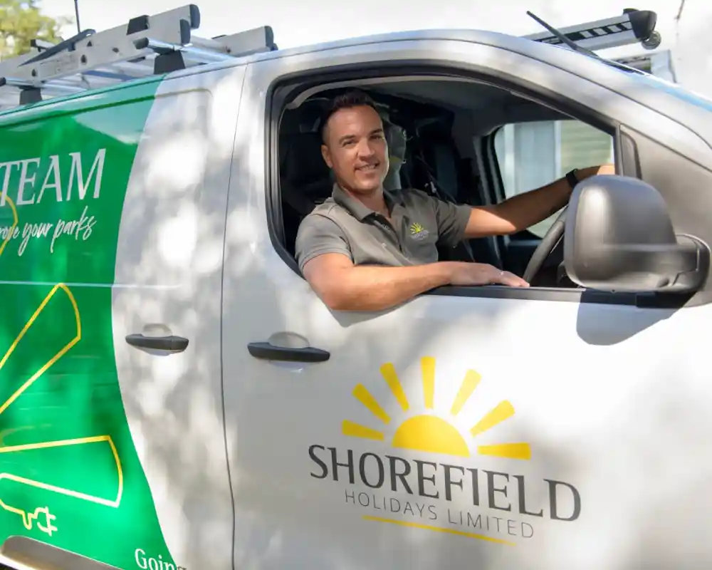 A man sits in the driver’s seat of a van branded with "Shorefield Holidays Limited," featuring a sun logo and green design elements. The man smiles and leans out the window, wearing a polo shirt with the company's logo.