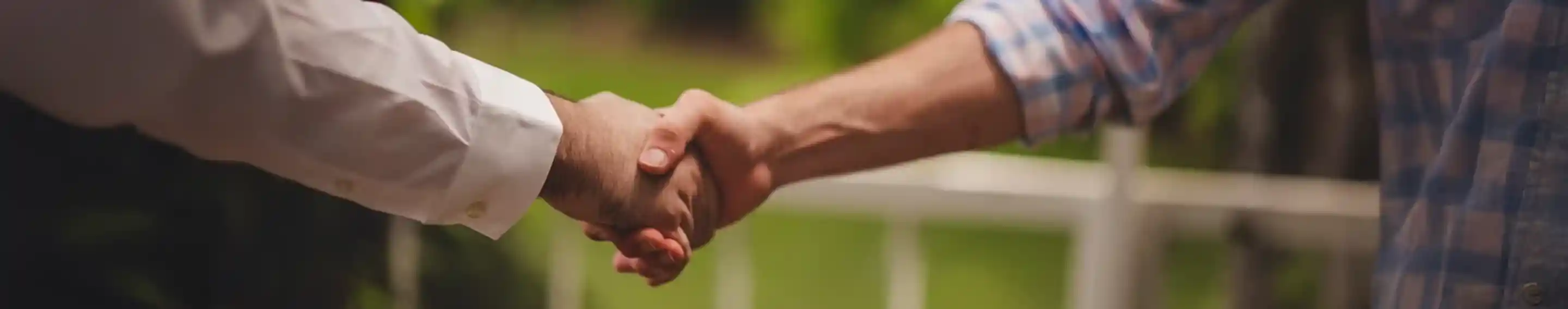 Two hands clasping in a handshake, set against a blurred green background. One hand is wearing a white long-sleeve shirt, and the other is in a blue and white checkered shirt.