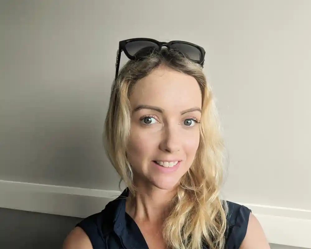 A woman with long, blonde hair and bright blue eyes smiles at the camera. She wears a dark top and has sunglasses resting on her head, with a neutral-colored wall in the background.