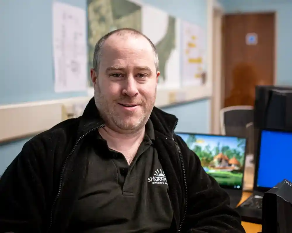A man with short hair and a slight beard smiles while seated at a desk in an office. He wears a black jacket with a logo. Behind him, there are charts and a computer displaying a scenic landscape.