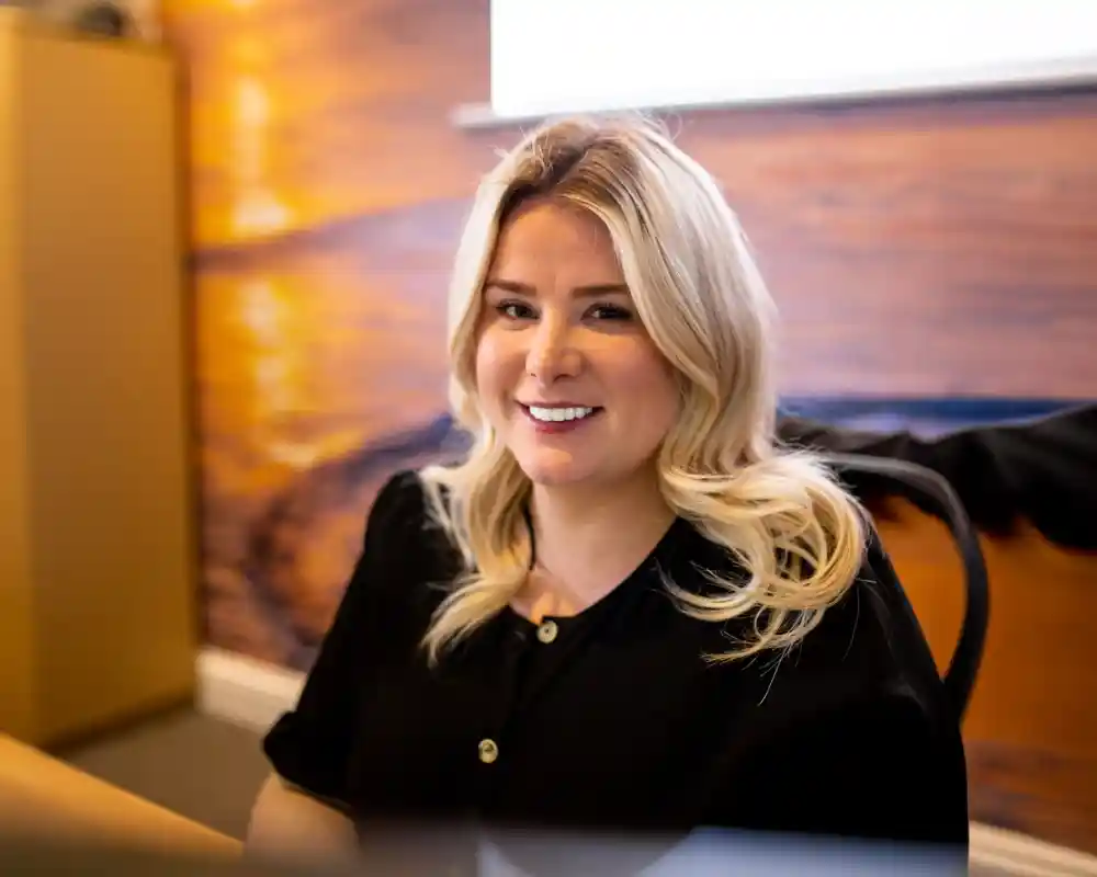 A smiling woman with long, blonde hair sits at a desk in an office. She is wearing a black blouse and appears to be engaged in a conversation or task. A warm, sunset-like mural is visible in the background.