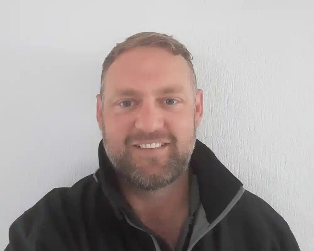A smiling man with short hair and a beard is wearing a black jacket. He is standing in front of a white textured wall.