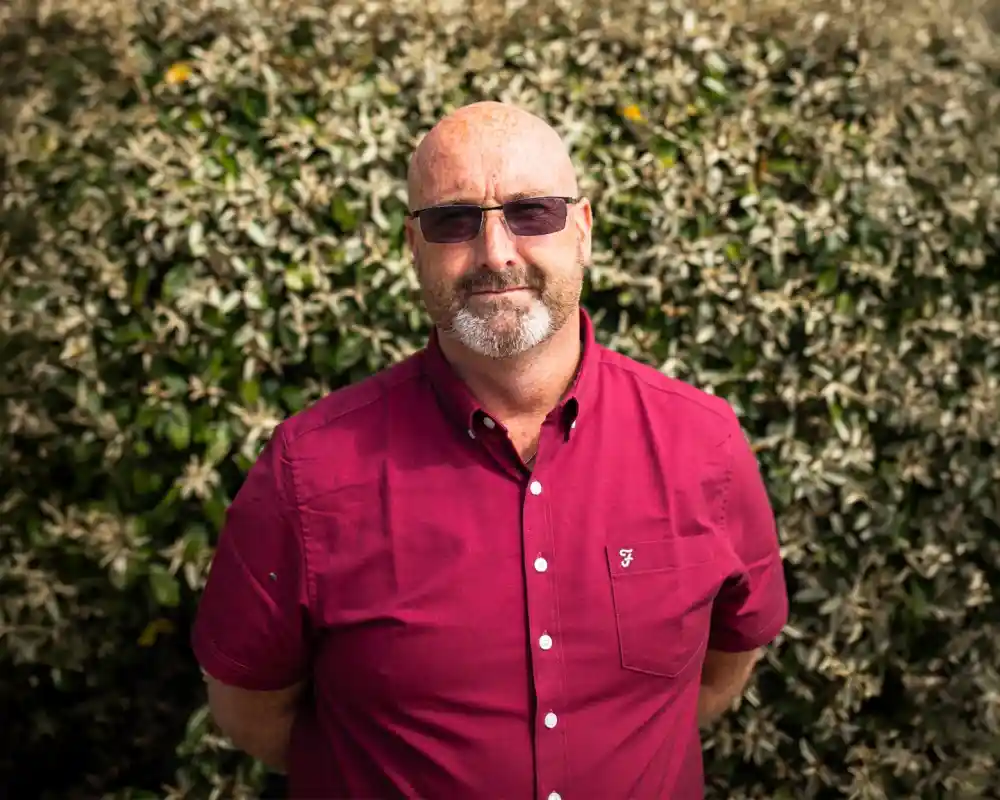A man with a bald head and salt-and-pepper beard stands in front of a leafy green background, wearing sunglasses and a maroon button-up shirt. His arms are crossed, and he has a serious expression.