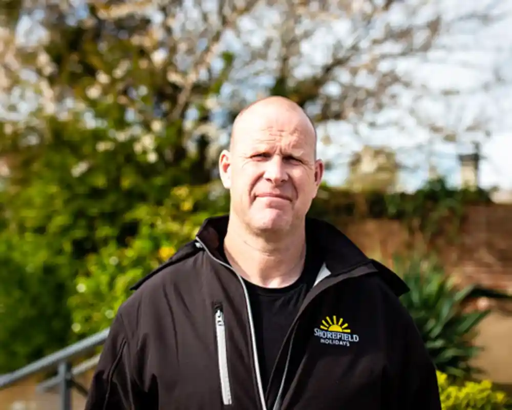 A man wearing a black jacket with a logo stands outside in a green, leafy environment. He has a serious expression and is looking directly at the camera. Trees and foliage are visible in the background under a partly cloudy sky.