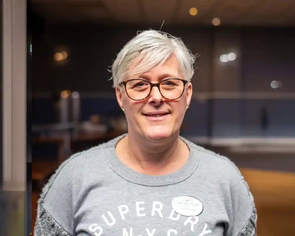 A woman with short gray hair and glasses smiles confidently while wearing a gray sweatshirt with the word "Superdry" printed on it. She stands in a softly lit indoor setting.