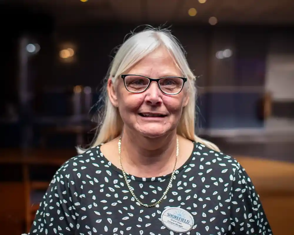 A woman with long, straight blonde hair wearing glasses and a patterned black shirt smiles at the camera. She has a name tag pinned to her shirt and is standing in a well-lit indoor environment.