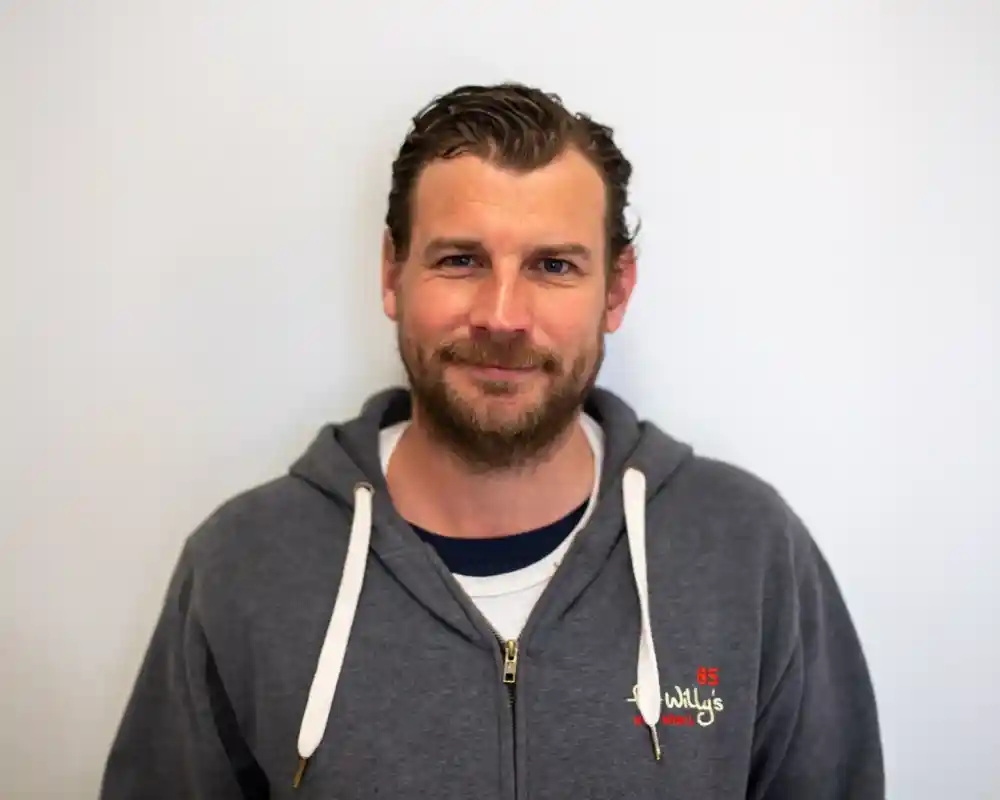 A man with dark hair and a beard smiles at the camera, wearing a gray hoodie. He is standing against a plain white background.