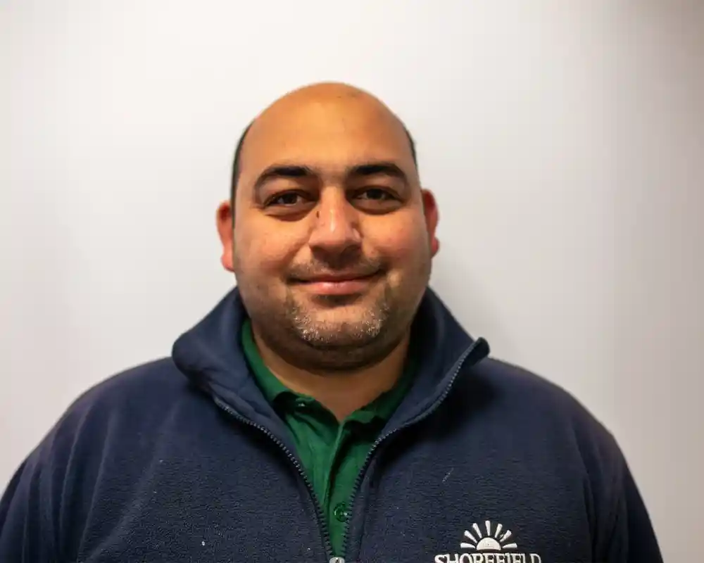 A man with a shaved head and light beard smiles at the camera, wearing a dark blue zip-up jacket over a green polo shirt. He stands against a plain white background.