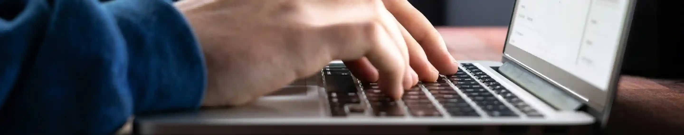 Person typing on a laptop at a wooden desk near a window, with natural light and greenery visible outside. The screen shows a web page with a form or document open.