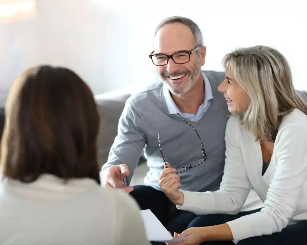 A smiling middle-aged man and woman engage in a conversation with another person, sitting together in a cozy living room. The man holds a pair of glasses, while the woman looks happily at him. All three appear to be in a relaxed and positive atmosphere.