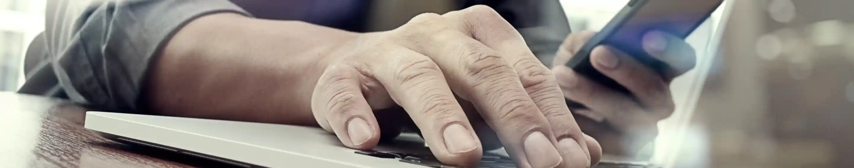 A person's hand typing on a laptop keyboard while holding a smartphone in the other hand. The scene is illuminated with soft light, creating a modern, dynamic atmosphere.