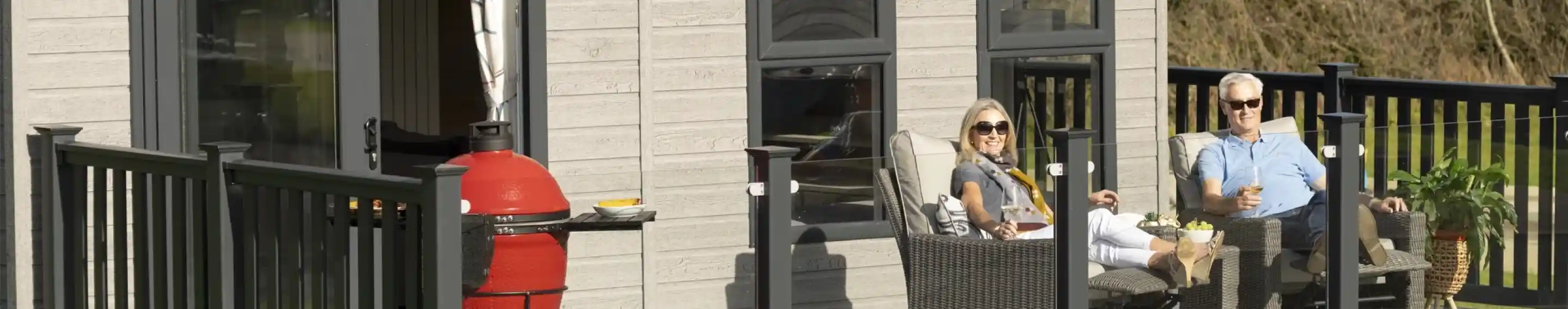 A couple relaxes on a modern deck of a cabin, enjoying drinks and sunshine. One person sits in a lounge chair, wearing sunglasses, while the other leans back with a relaxed posture. A red grill is visible nearby, and greenery surrounds the cabin.