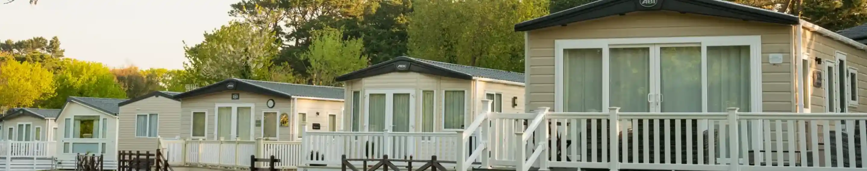 Row of holiday cabins with white railings, set on a grassy area under a blue sky. Lush green trees and shrubs are in the background.