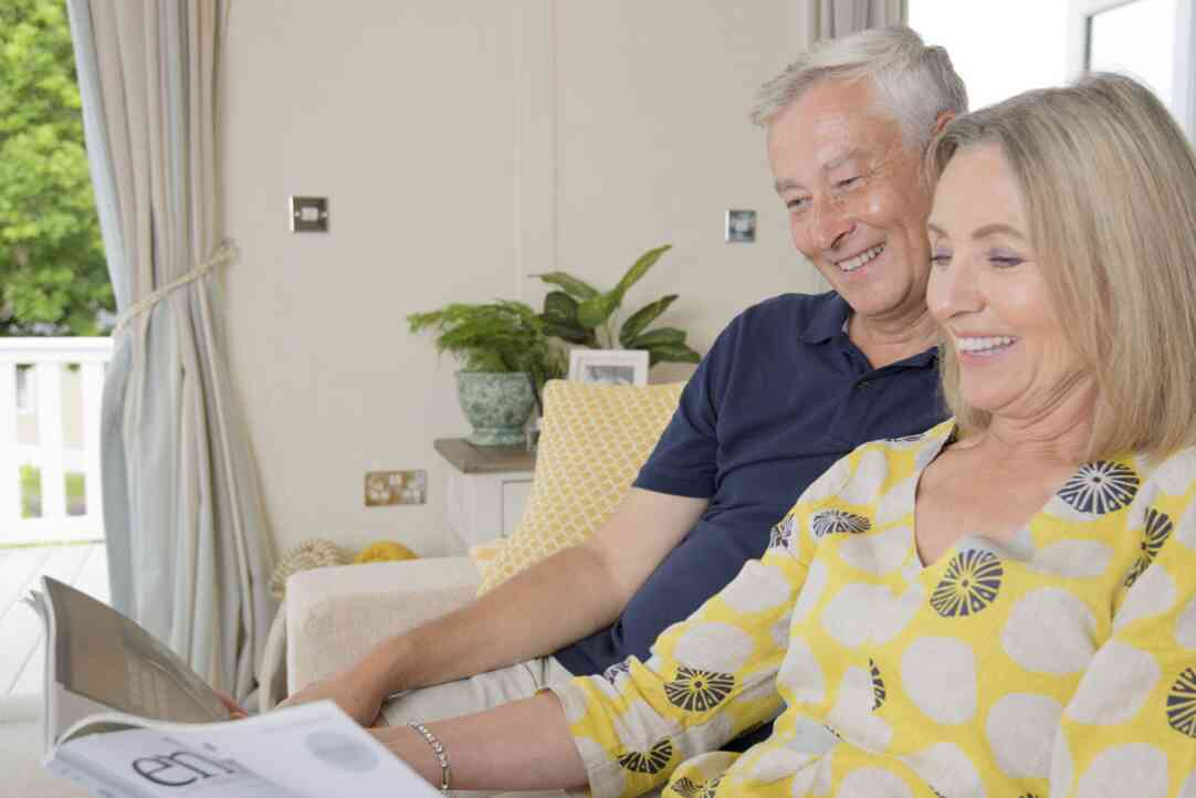 A senior couple sits comfortably on a couch, engaged in reading a magazine, smiling at each other in a homely setting.