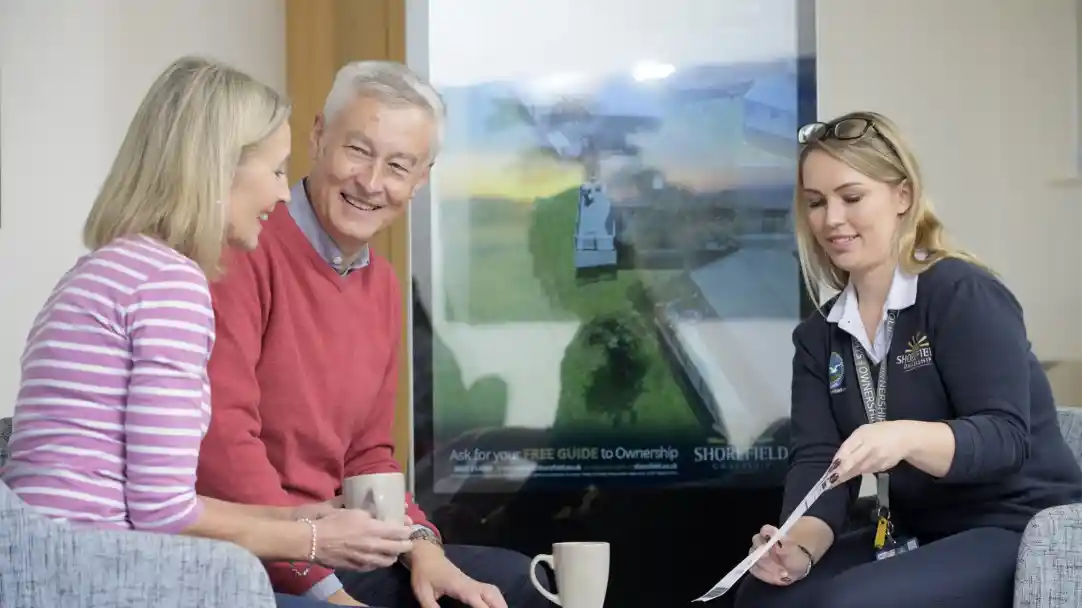 A man and woman sit together, smiling and talking with a woman in a dark uniform who holds a brochure. A large screen in the background displays an aerial view of a property.