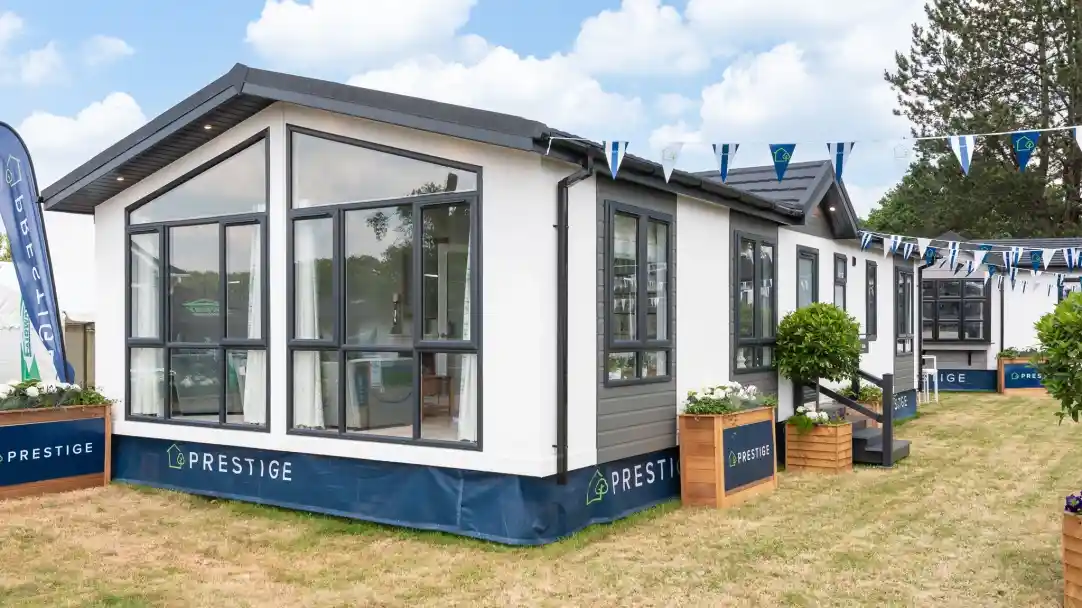 A modern prefabricated home with large windows, situated on grassy land. Colorful bunting hangs above, and landscaping features potted plants. The home has a sleek design with a black and white exterior.