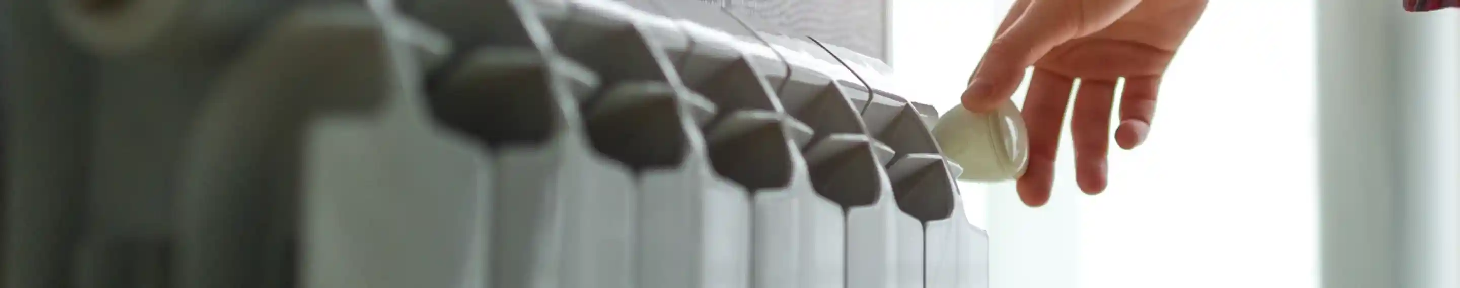 A hand is adjusting the temperature knob on a radiator, with soft natural light filtering through a window in the background.