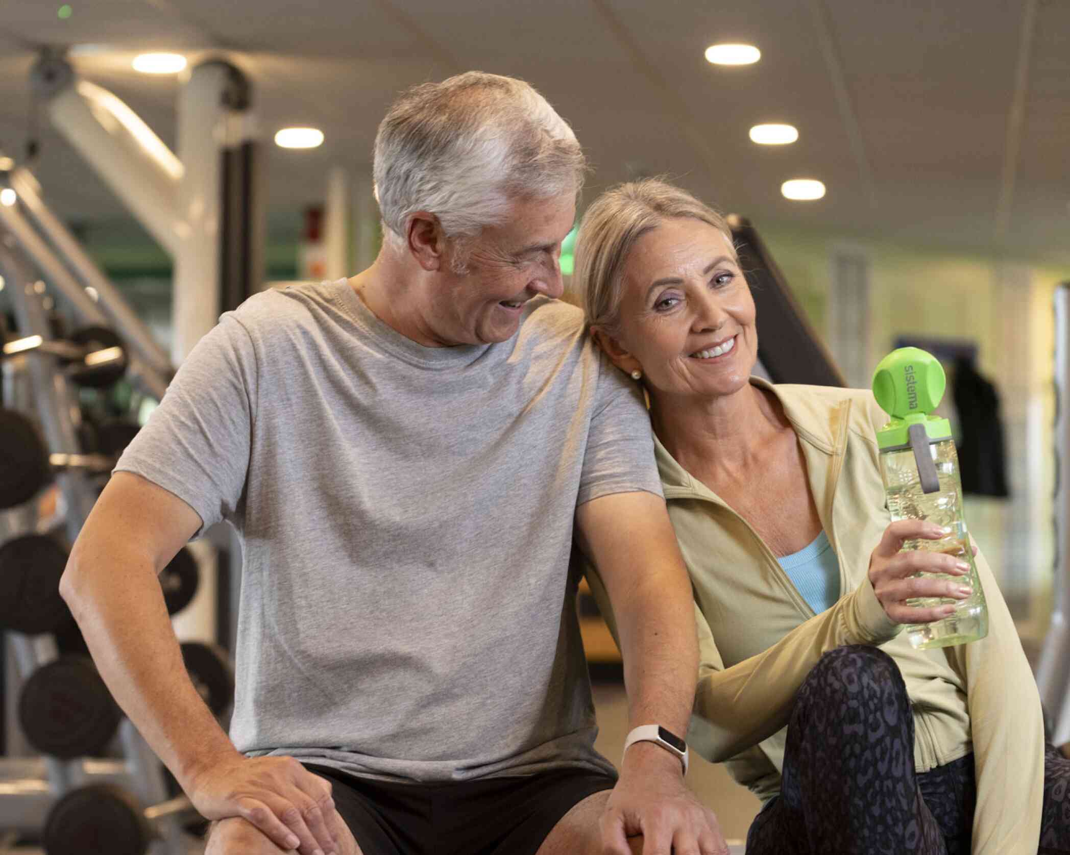 A smiling older couple sits together in a gym, with the woman holding a green water bottle. They appear to be enjoying each other's company after a workout. The gym features exercise equipment in the background.