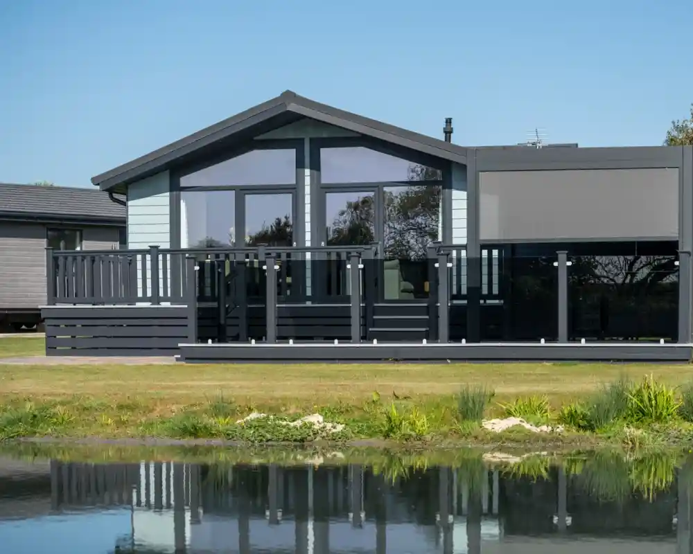 A modern house with large windows and a black deck overlooks a calm pond surrounded by greenery. The sky is clear and blue.