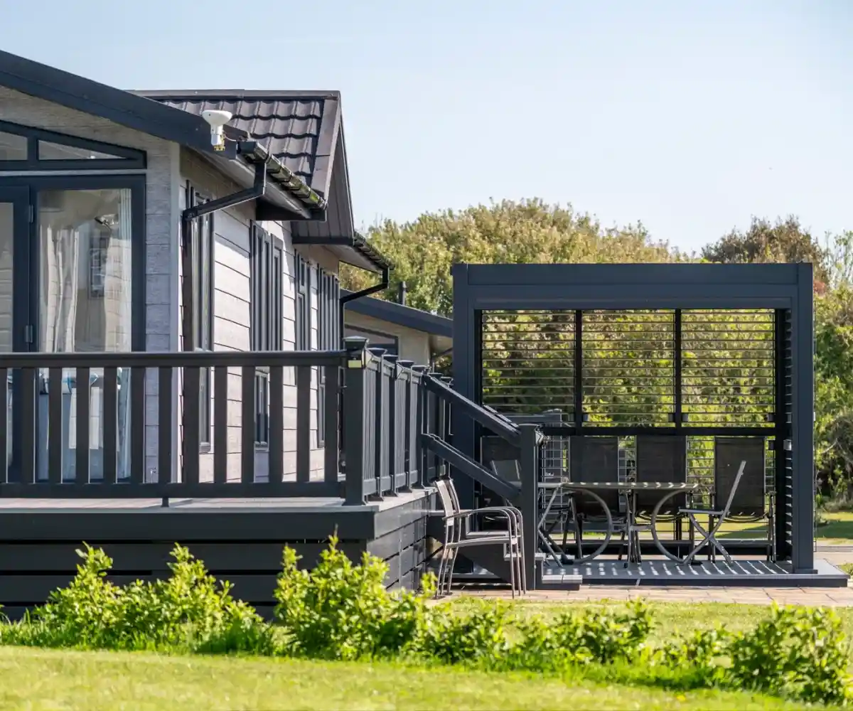 A modern wooden cabin is situated on a grassy hillside, surrounded by tall trees. The cabin features a spacious deck and large windows, offering a serene outdoor view.