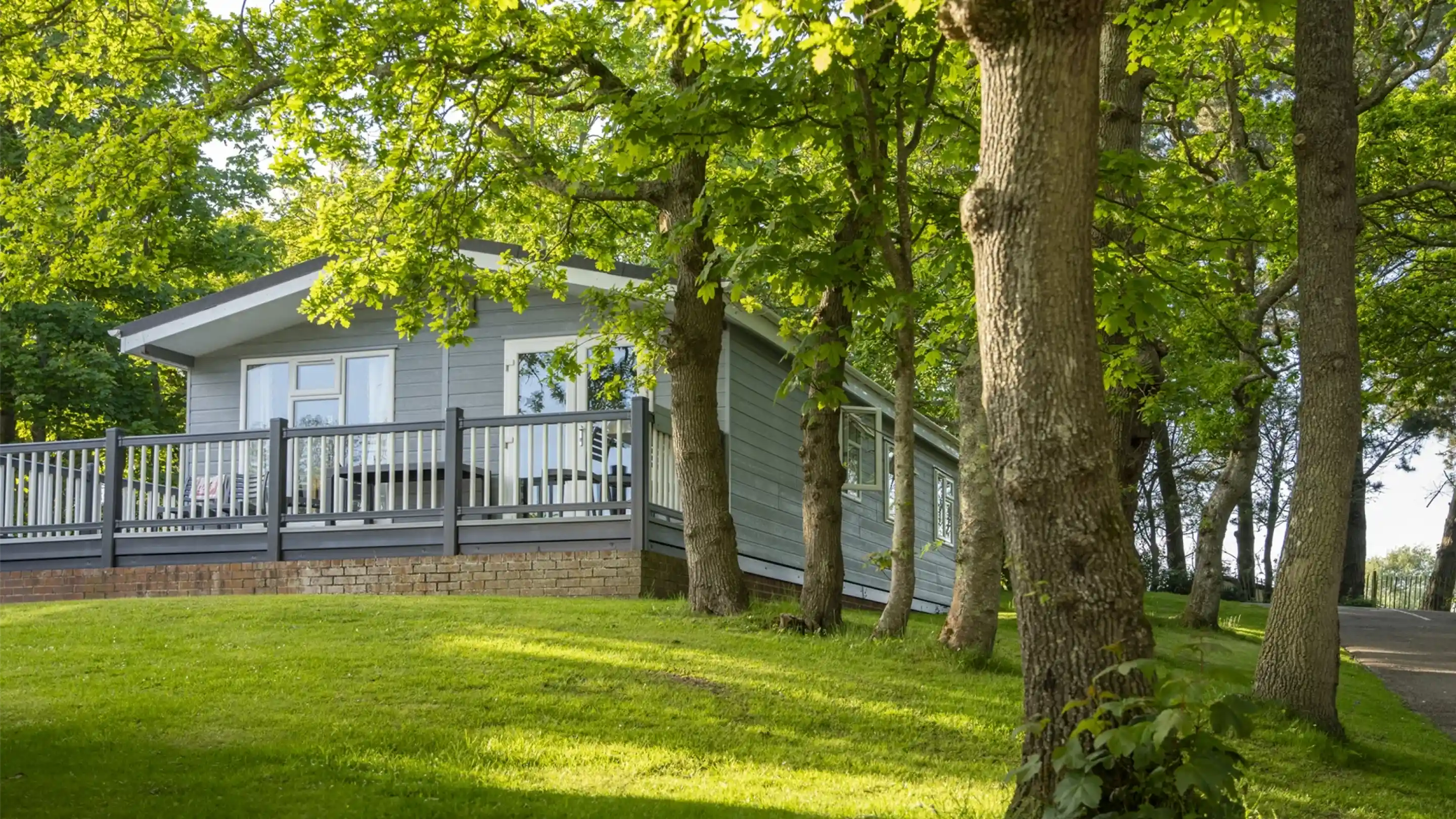 A modern wooden cabin is situated on a grassy hillside, surrounded by tall trees. The cabin features a spacious deck and large windows, offering a serene outdoor view.