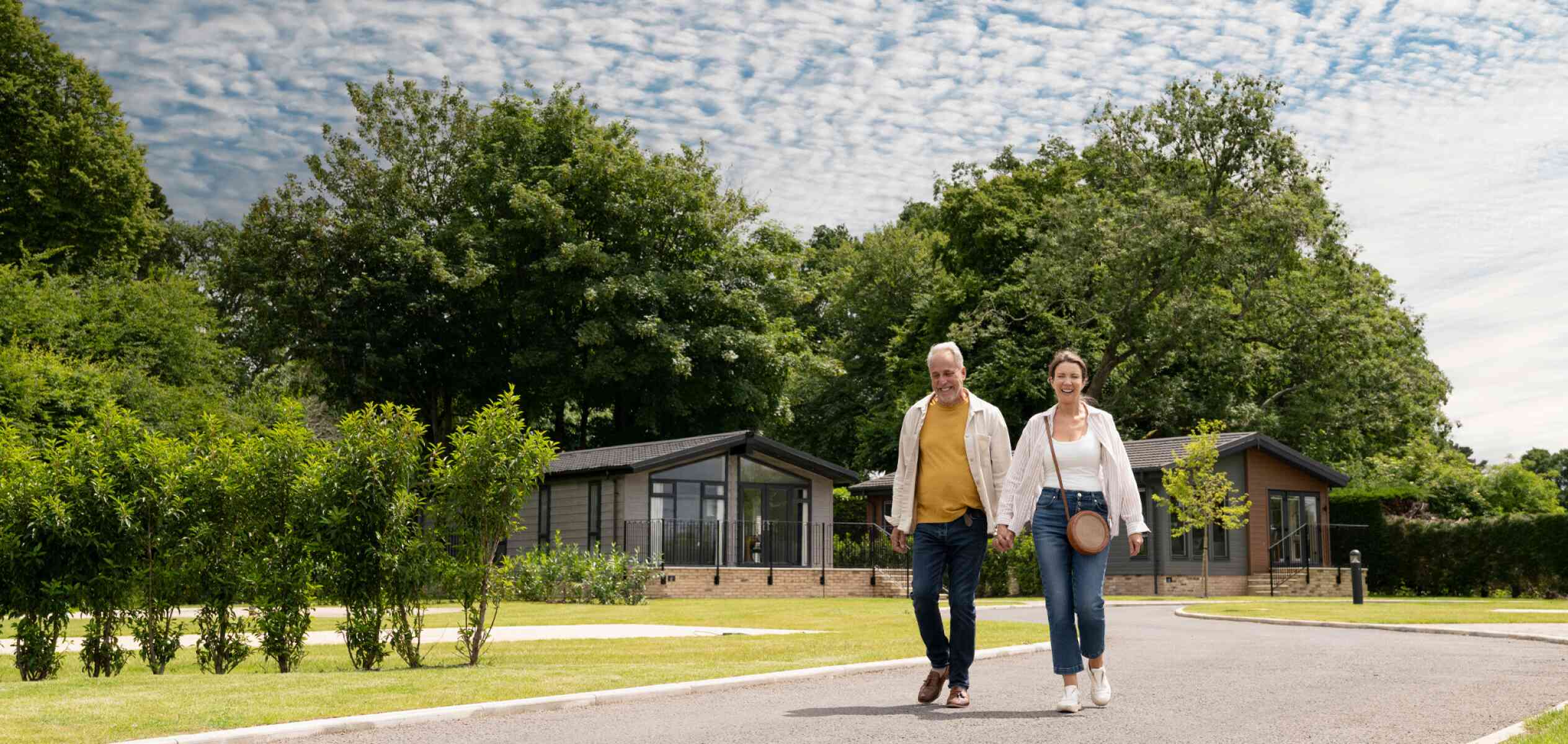 An older man and a woman walk down a tree-lined pathway, smiling and holding a small ball. Modern cabins are visible in the background, set against a bright blue sky with scattered clouds.
