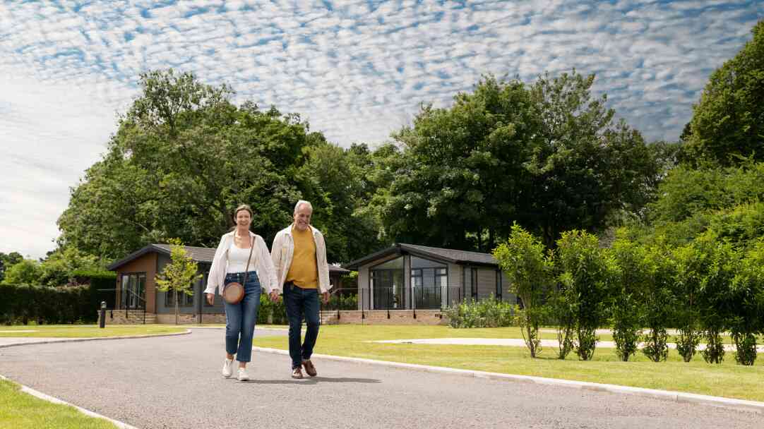 A middle-aged couple walks hand in hand along a pathway in a scenic park, surrounded by lush greenery and modern holiday cabins in the background. The sky is partly cloudy.
