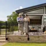 A man and a woman stand on a balcony, enjoying drinks and conversing. The woman wears a light-colored dress, while the man is in a shirt and beige pants. The background features a modern house with open doors and a patio area. Lush green grass is visible in front of them.