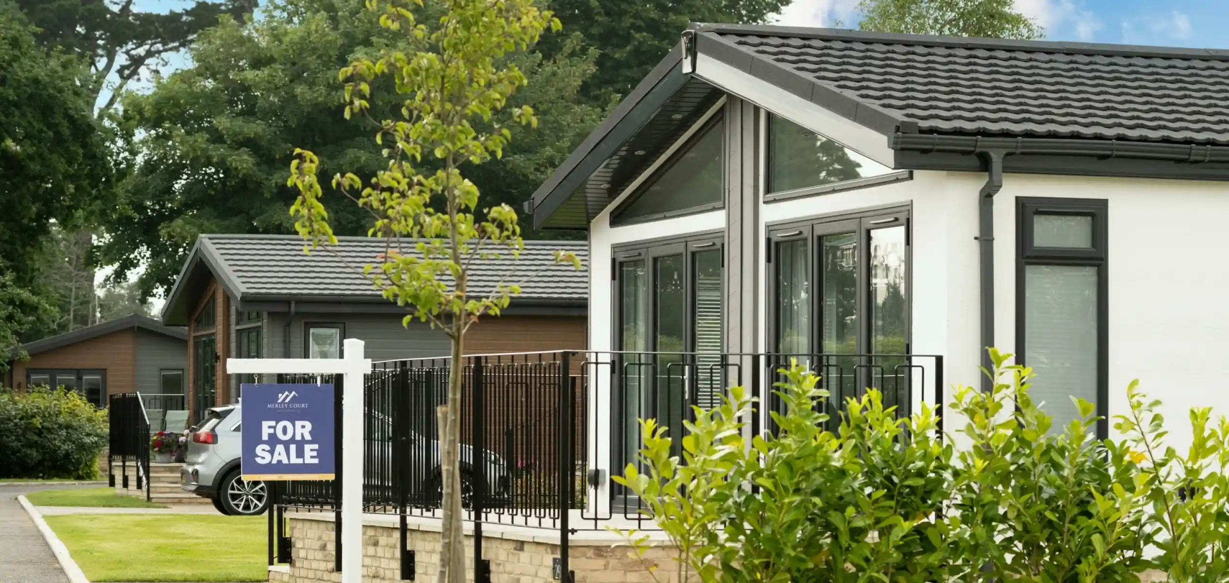 Modern homes for sale in a residential area, featuring a landscaped front with a "For Sale" sign. Lush greenery and trees surround the properties, creating a peaceful environment.