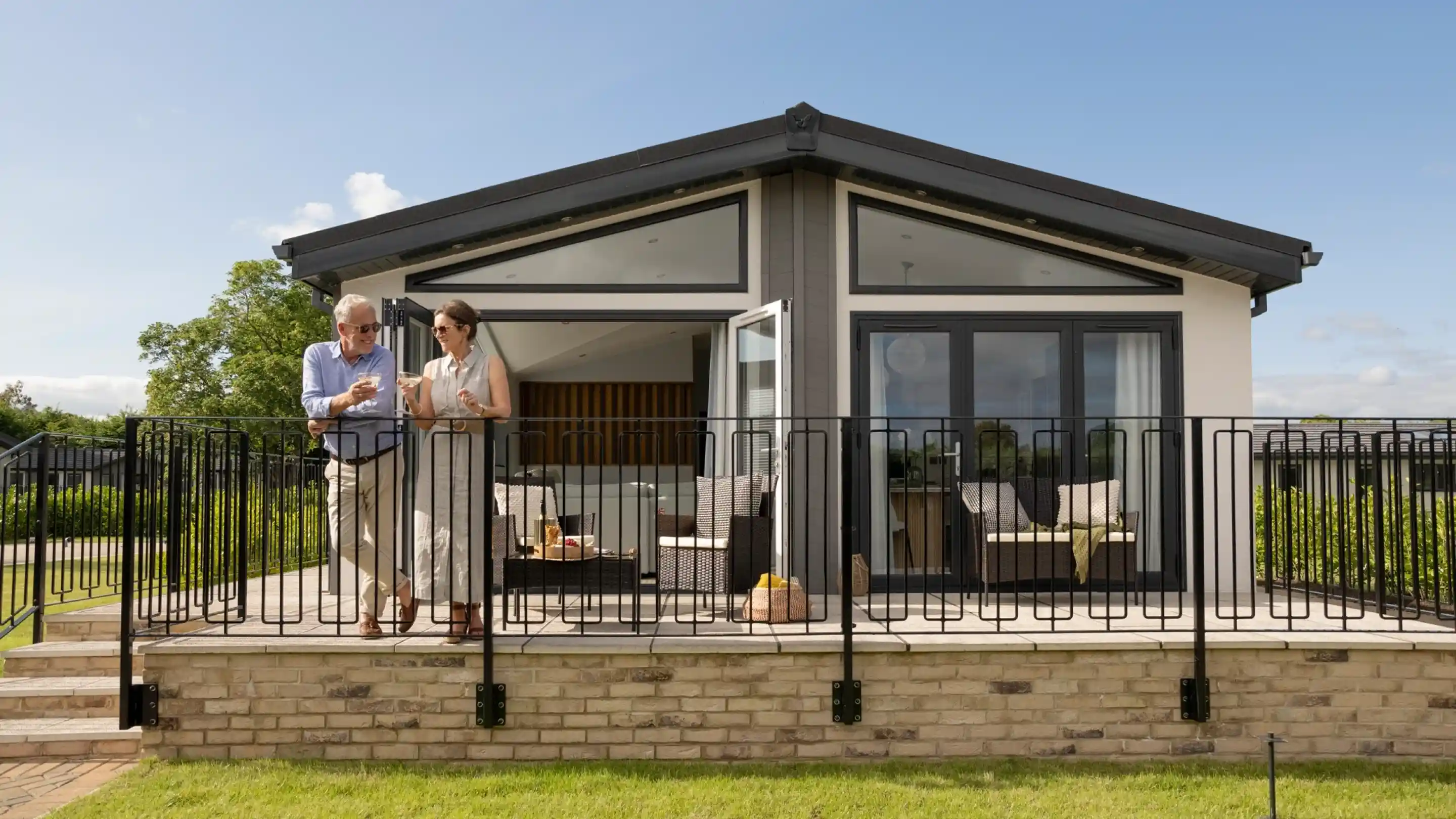A couple stands on a patio in front of a modern house, discussing something while enjoying a sunny day. The house features large windows and a contemporary design, with a green lawn in front.