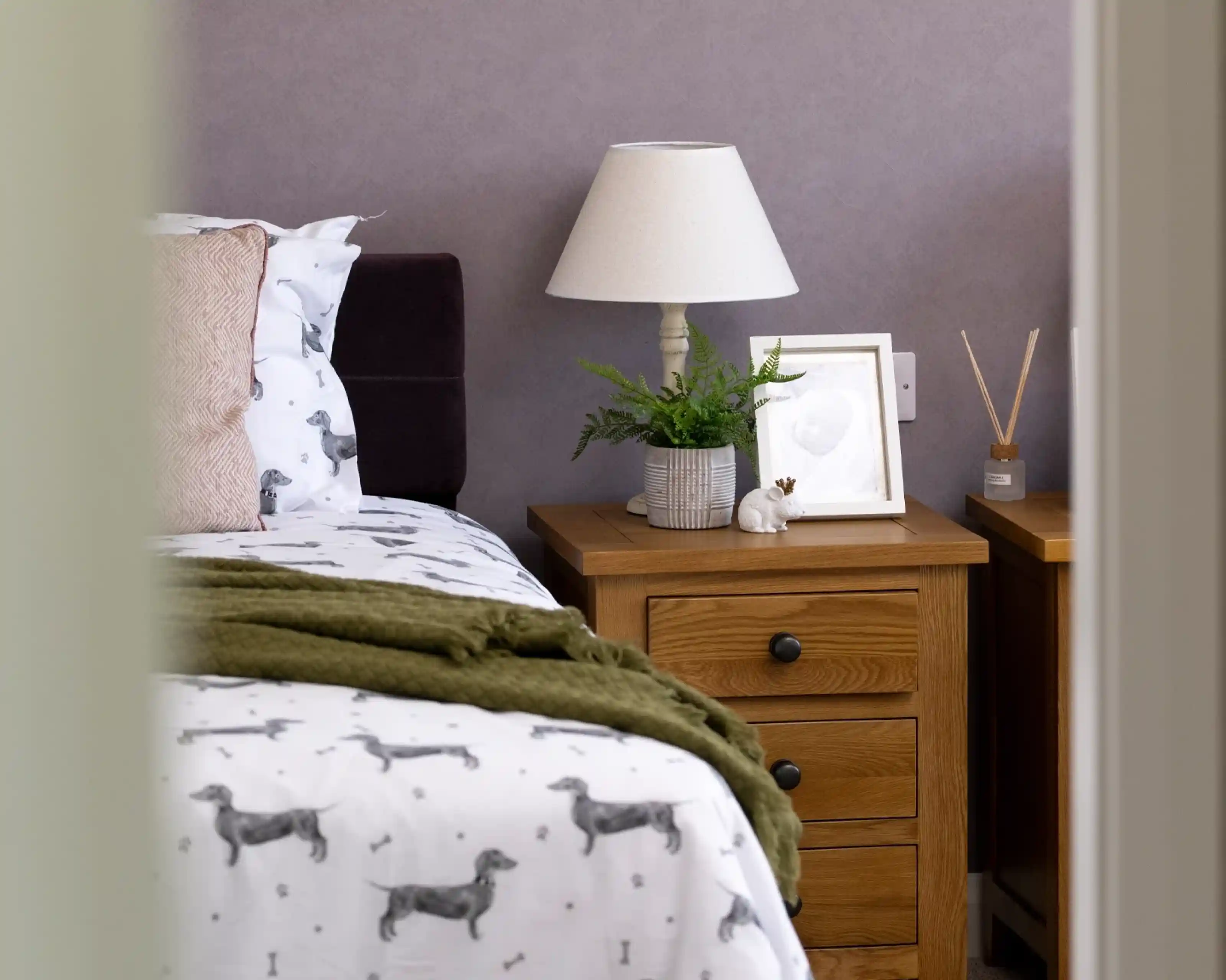 A cozy bedroom featuring a bed with dog-patterned white sheets, a bedside table with a lamp, a small potted plant, a framed picture, and a decorative rabbit. Walls are painted in a soft purple hue.