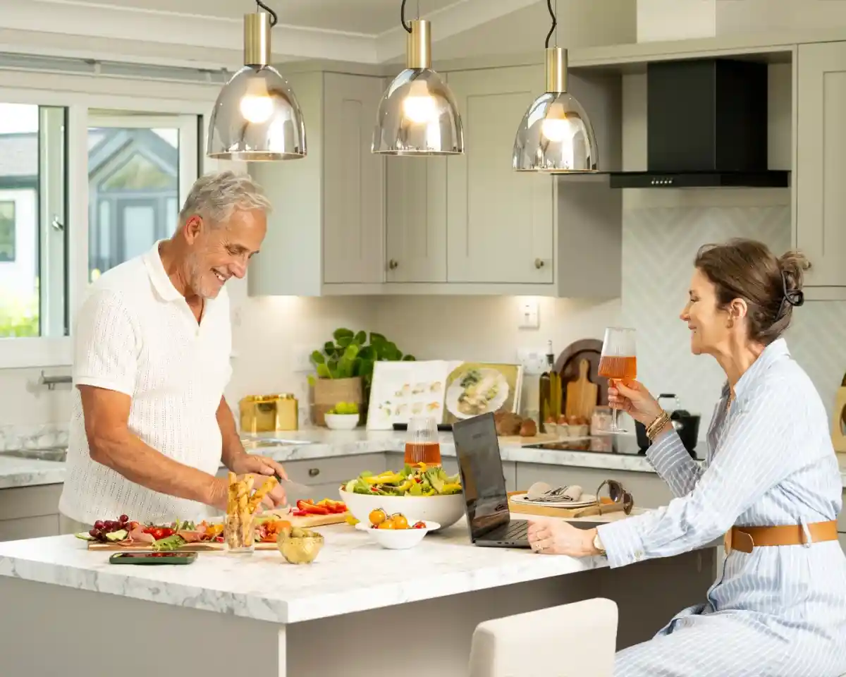 A man in a white sweater smiles while preparing food on a kitchen countertop. A woman in a striped dress sits at the table, holding a drink and looking at a laptop. Fresh fruits and salads are arranged on the counter, with bright pendant lights illuminating the cozy kitchen.