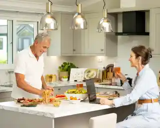 A man in a white sweater smiles while preparing food on a kitchen countertop. A woman in a striped dress sits at the table, holding a drink and looking at a laptop. Fresh fruits and salads are arranged on the counter, with bright pendant lights illuminating the cozy kitchen.