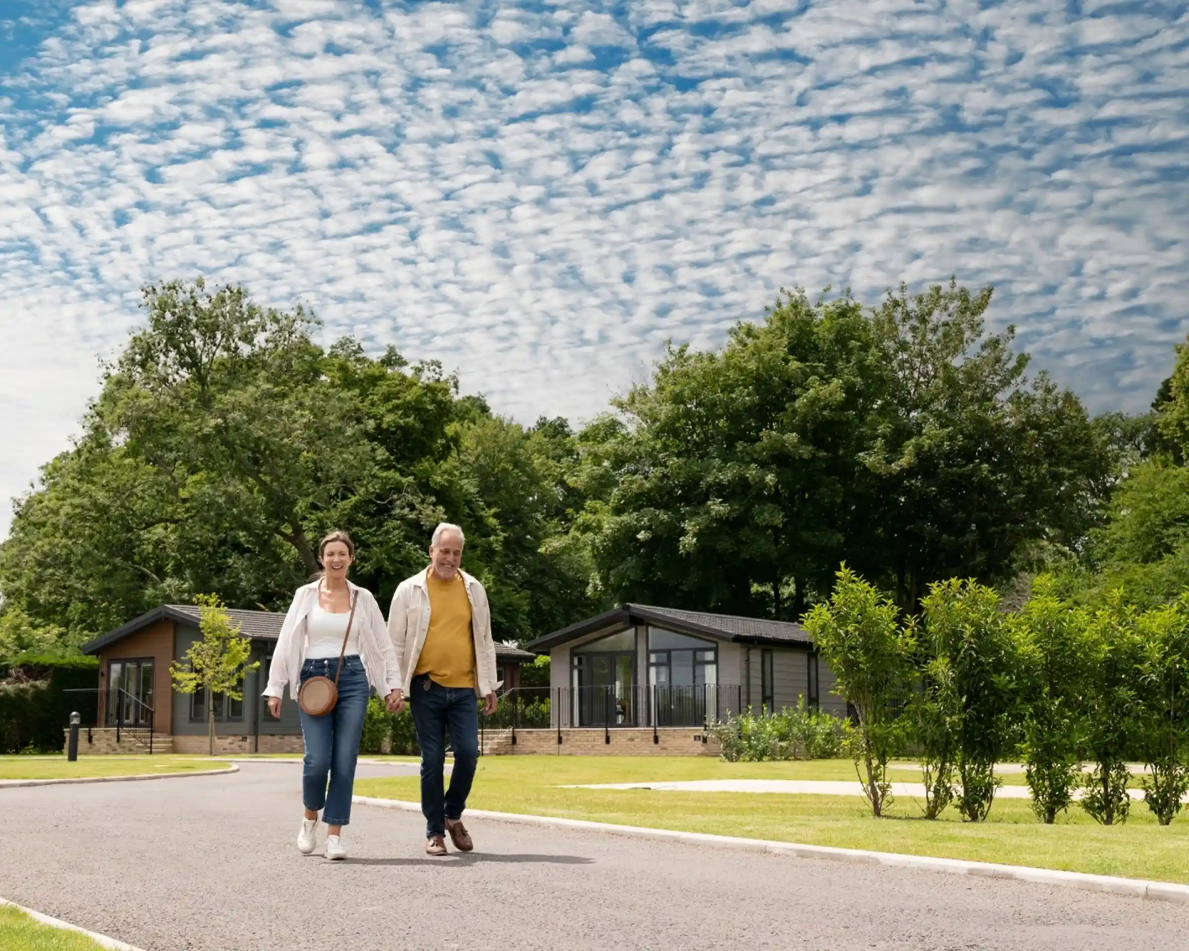 A cheerful older couple strolls hand in hand along a pathway lined with greenery and modern cabins under a cloudy blue sky.