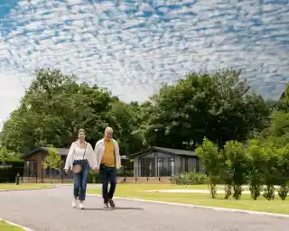 A cheerful older couple strolls hand in hand along a pathway lined with greenery and modern cabins under a cloudy blue sky.