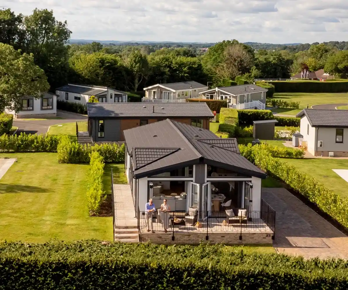 A modern wooden cabin is situated on a grassy hillside, surrounded by tall trees. The cabin features a spacious deck and large windows, offering a serene outdoor view.