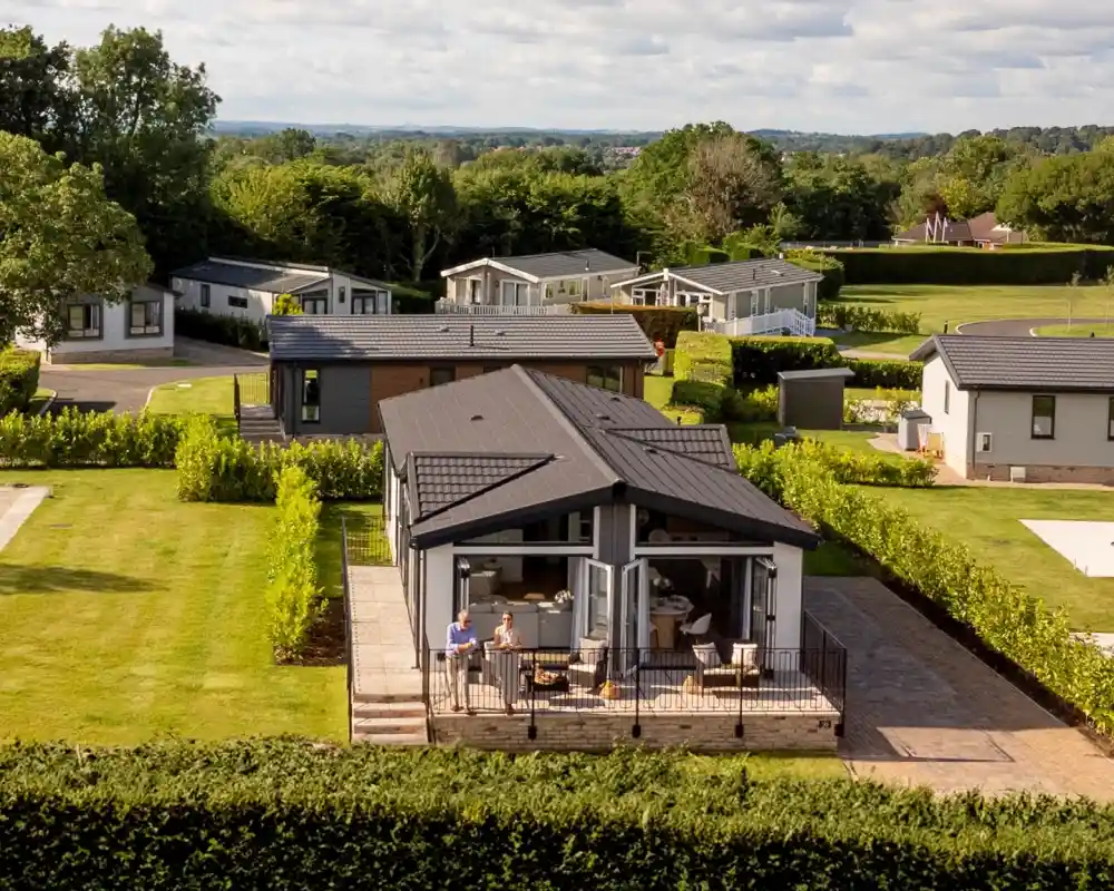 A modern house with a dark roof sits in a landscaped area, surrounded by lush greenery. Two people are enjoying a meal on the patio. Nearby, several similar homes are visible, set against a backdrop of trees and a cloudy sky.