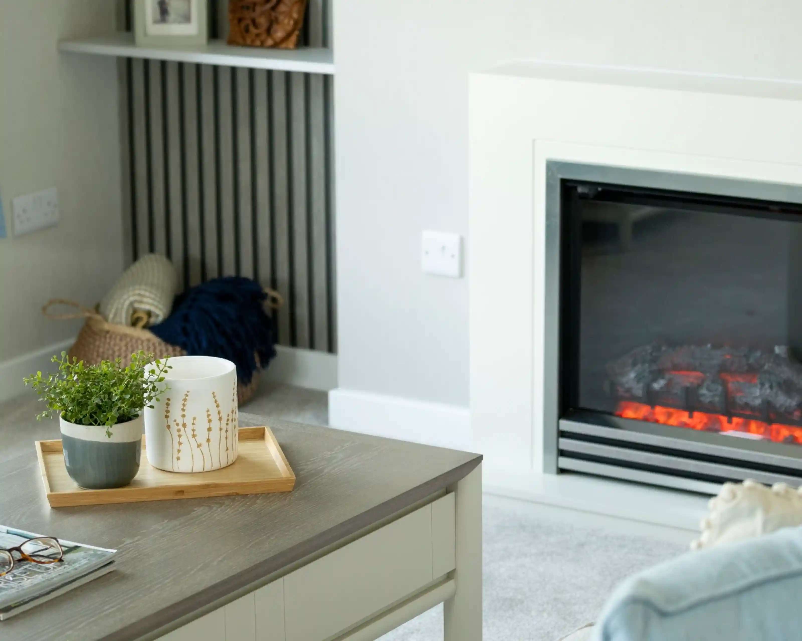 A cozy living room featuring a stylish table with a wooden tray holding a potted plant and a decorative candle. In the background, a modern fireplace emits a warm glow, while a blue throw and a basket add texture to the space.