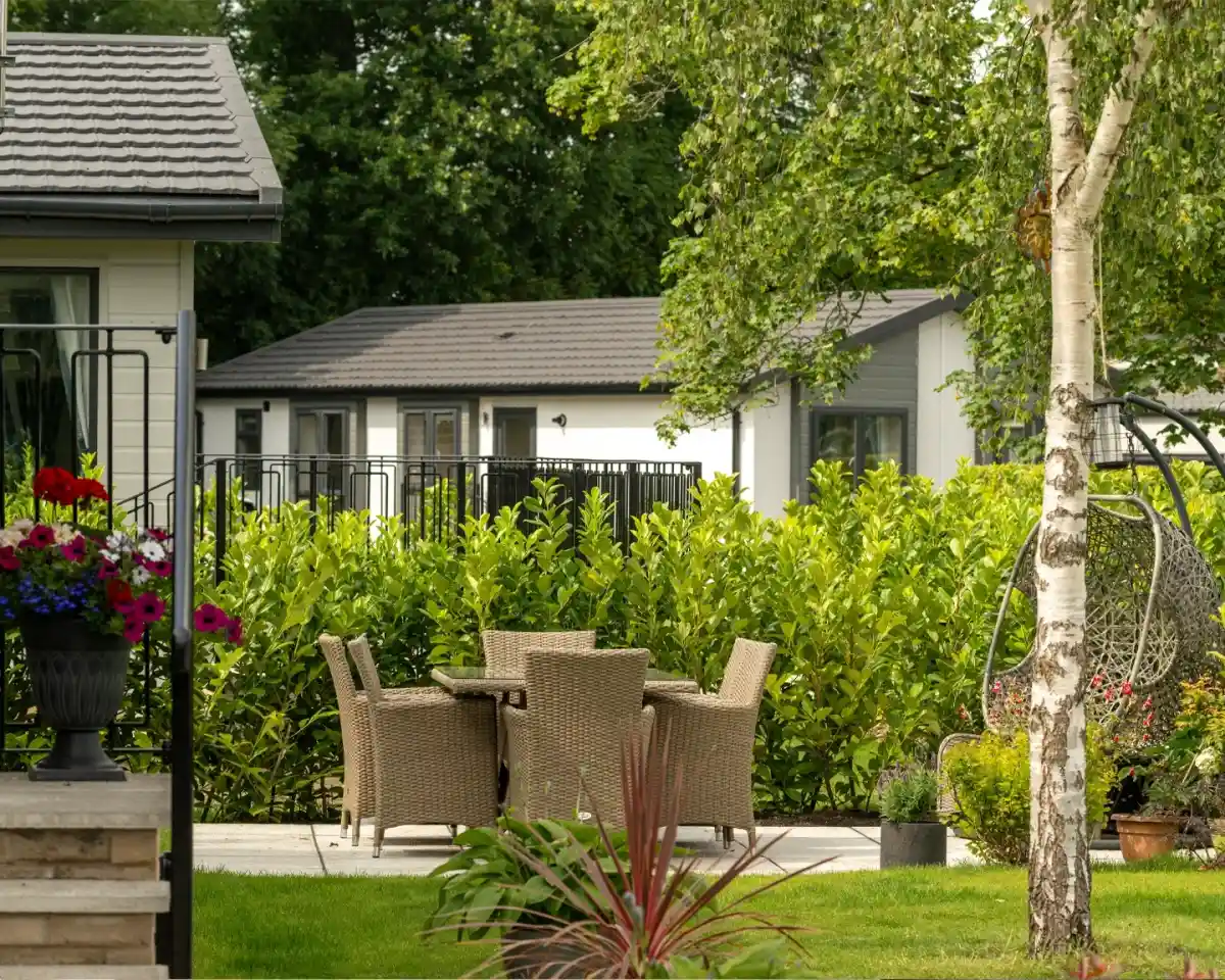 A cozy outdoor patio featuring a woven dining set surrounded by lush green hedges and a birch tree. In the background, modern homes are partially visible. Bright flowers are in pots near the patio area.