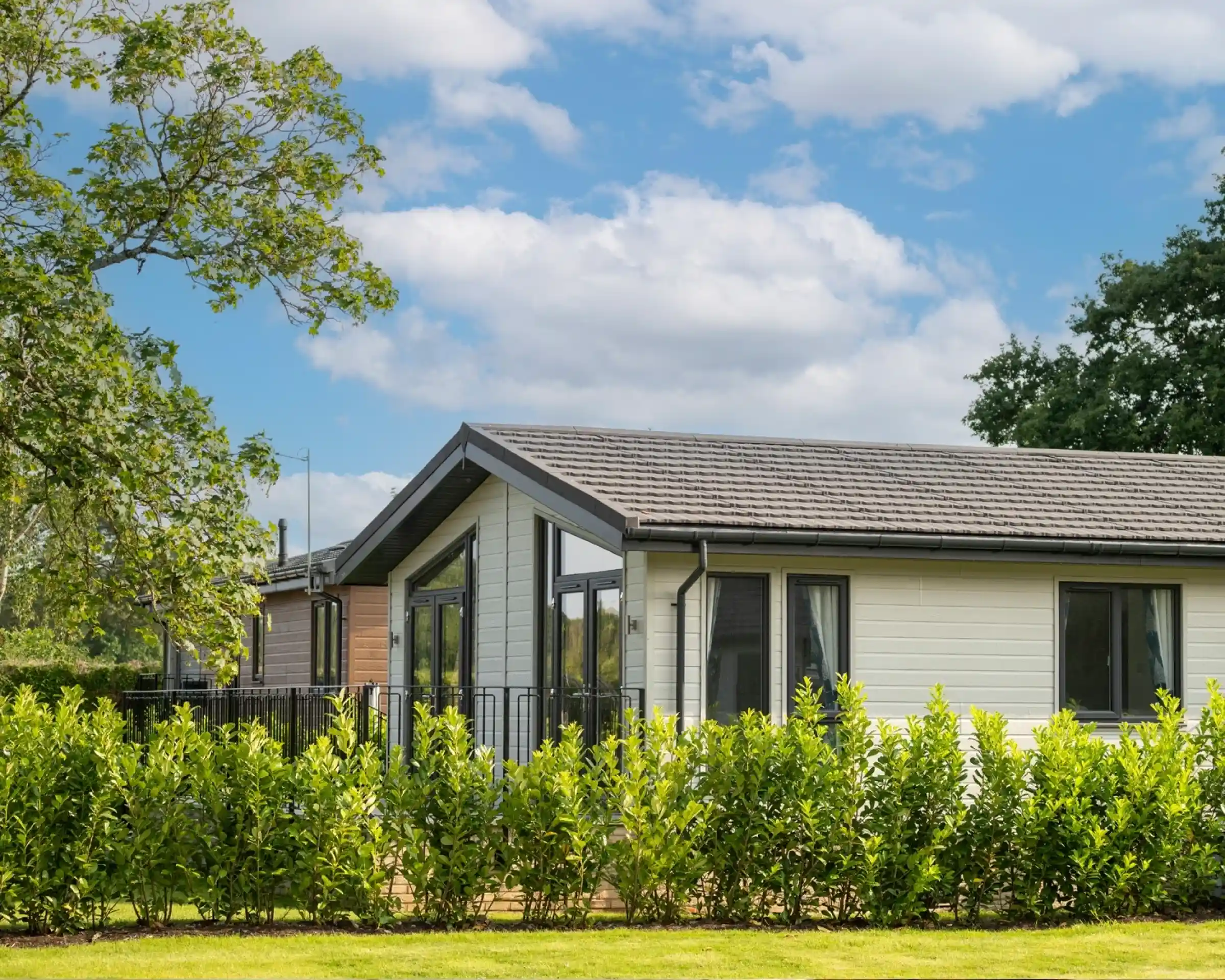 A modern single-story house with a gray roof and large windows, surrounded by a green hedge and lush grass under a partly cloudy sky.