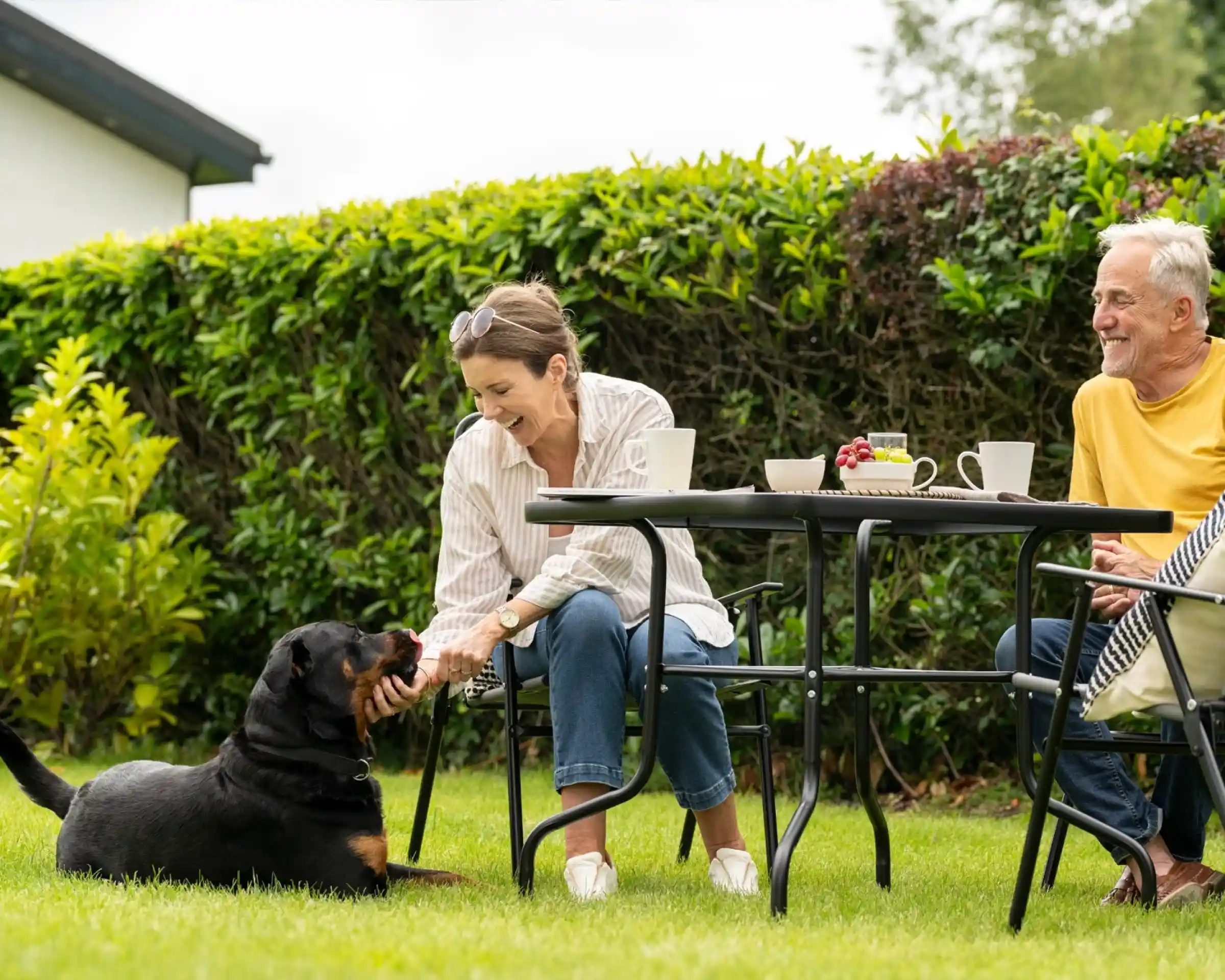 A woman sits at a garden table, smiling and petting a black dog. An older man sits nearby, enjoying a cup of coffee. Green bushes surround the scene, and a few snacks are on the table.