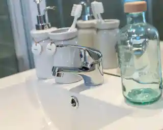 A modern bathroom sink with a sleek chrome faucet. Nearby are two white containers holding toothbrushes and other accessories, and a glass bottle with a wooden cap. The countertop is smooth and white, reflecting a clean and organized space.