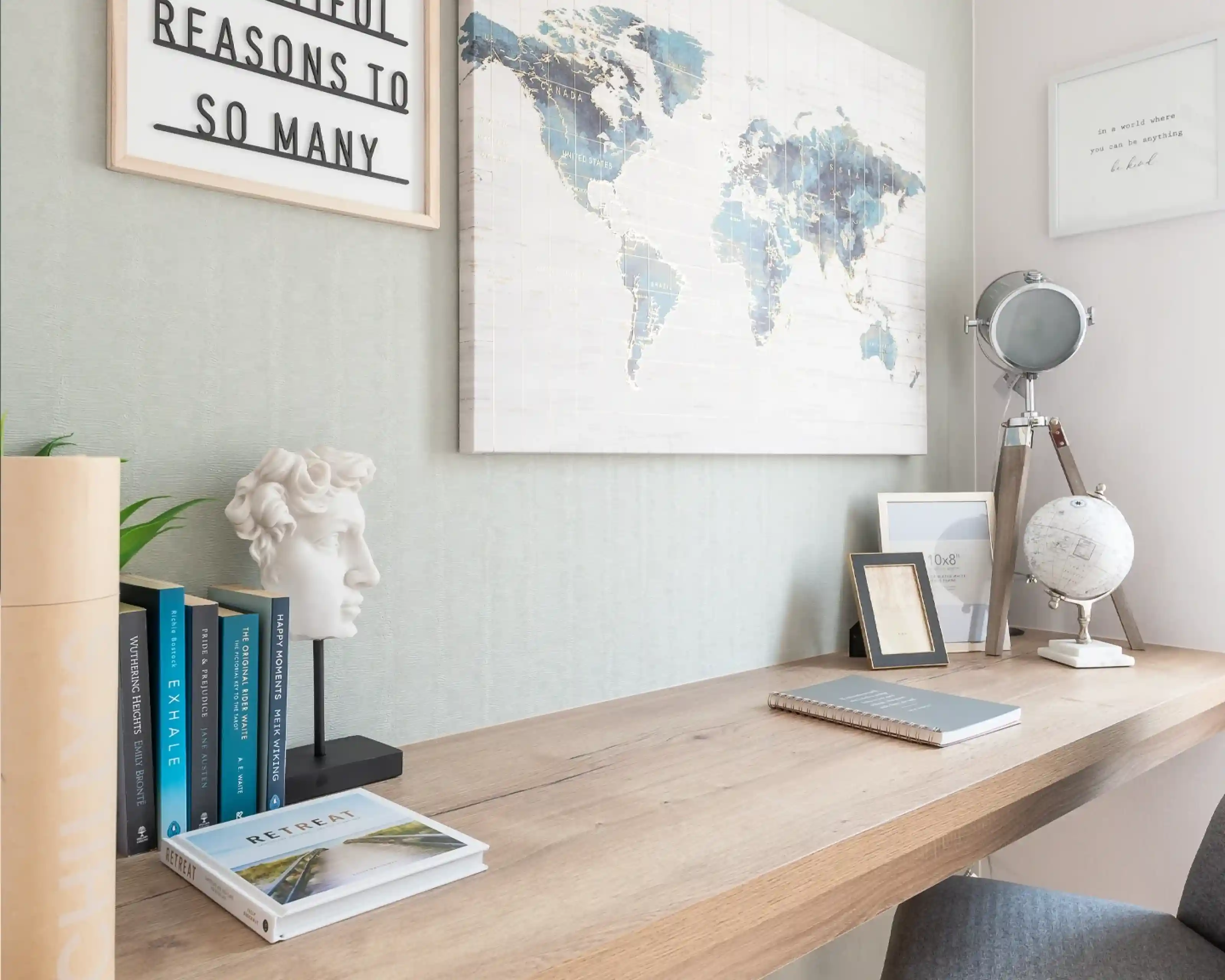 A modern workspace featuring a wooden desk with books, a small globe, a framed quote, and a decorative bust. A world map hangs on the wall, complementing the serene color palette of the room.