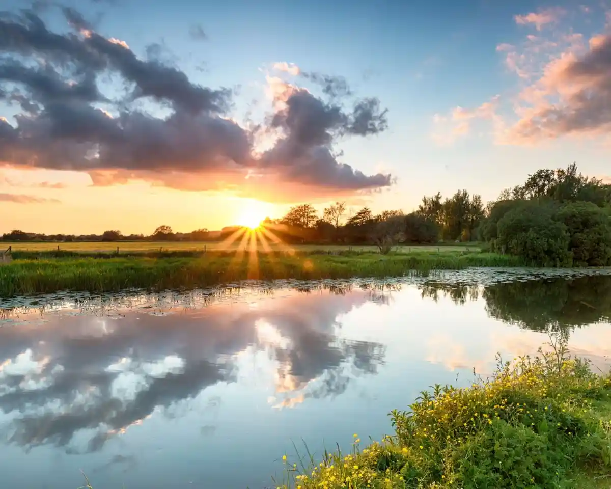 Sunset over a tranquil river, with vibrant orange and pink clouds reflecting on the water. Lush greenery and trees line the banks, creating a serene landscape.