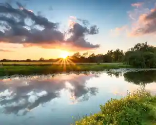 Sunset over a tranquil river, with vibrant orange and pink clouds reflecting on the water. Lush greenery and trees line the banks, creating a serene landscape.