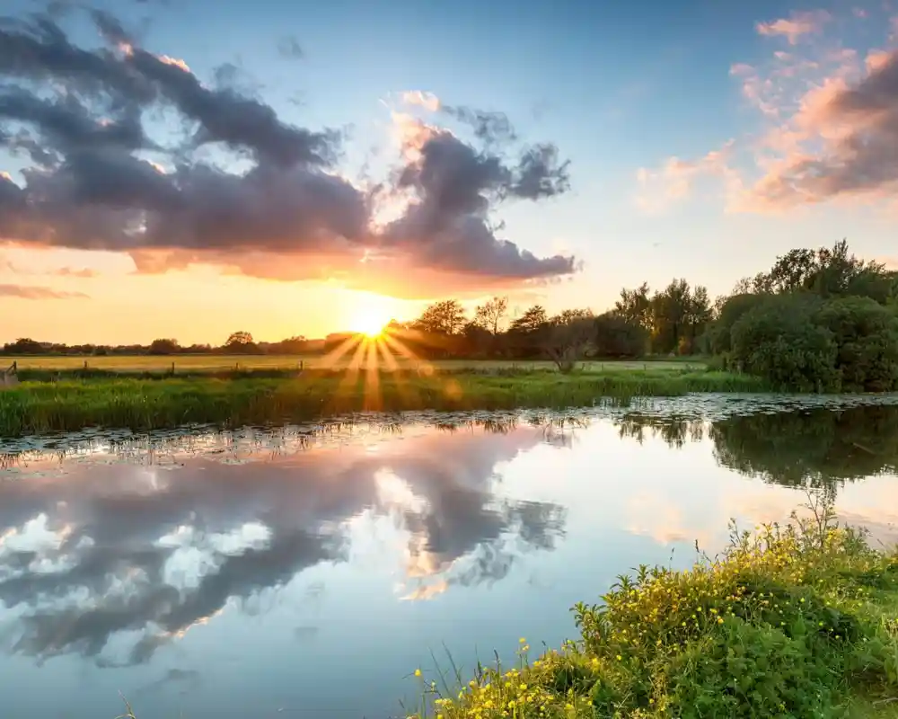 Sunset over a tranquil river, with vibrant orange and pink clouds reflecting on the water. Lush greenery and trees line the banks, creating a serene landscape.
