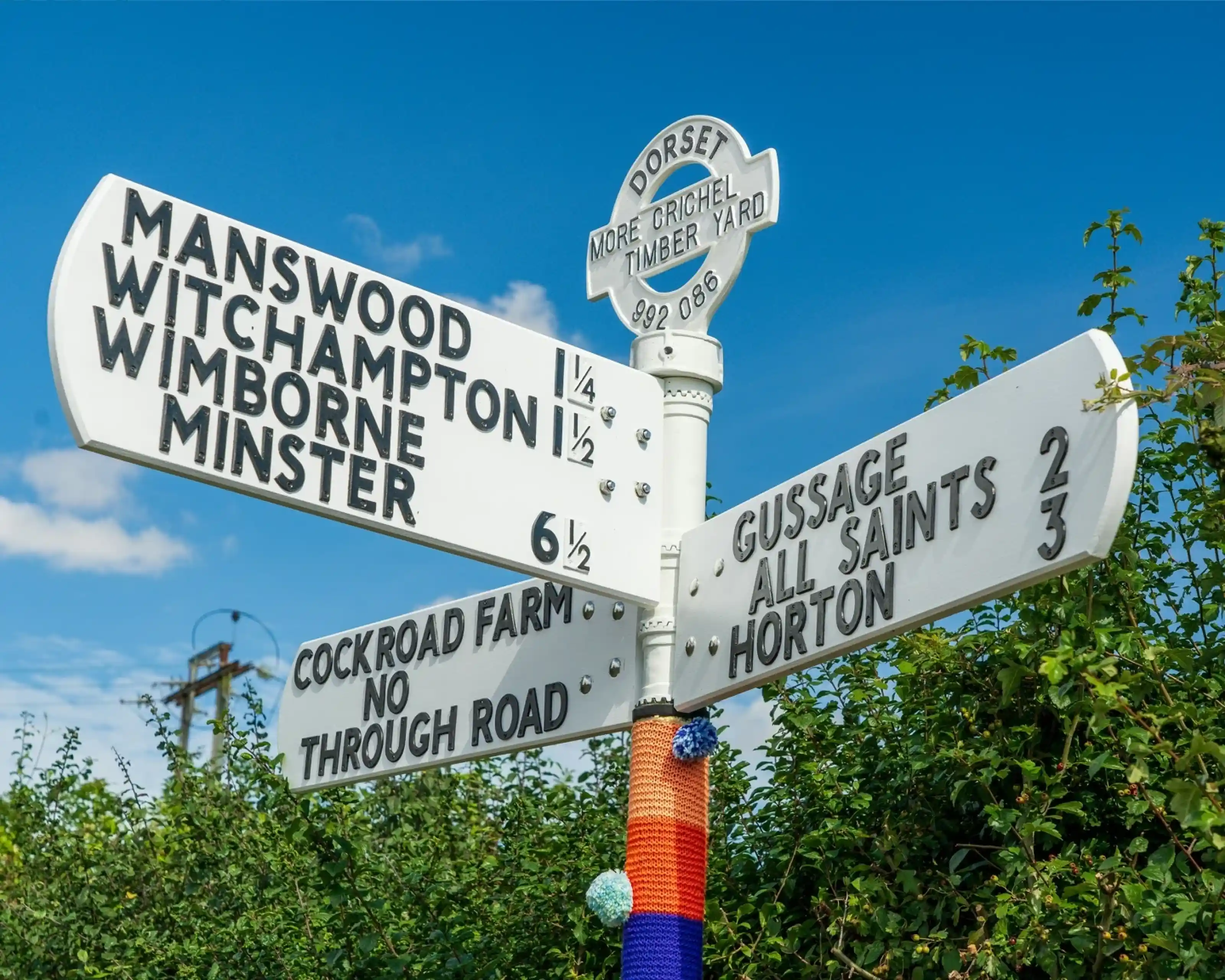 Directional signpost in Dorset showing distances to Manswood, Witchampton, Wimborne Minster, Gussage, All Saints, and Horton against a bright blue sky.
