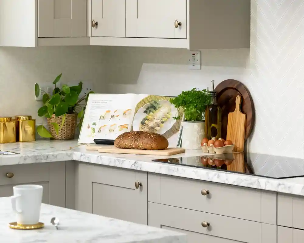 A cozy kitchen corner featuring a marble countertop, a loaf of bread on a wooden cutting board, a cookbook opened to a recipe, a small potted plant, fresh herbs in a bowl, and some eggs in a carton, with stylish cabinets and a backsplash in the background.
