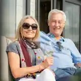 A smiling couple sits together outdoors, enjoying a sunny day. The woman, wearing sunglasses and a colourful scarf, holds a glass of white wine, while the man, in a light blue polo shirt, smiles next to her. They are seated on a comfortable couch with large windows in the background.
